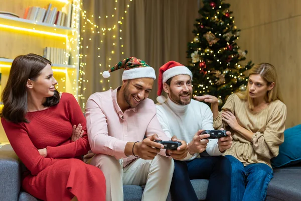 New Years party group of four diverse friends having fun relaxing and celebrating on Christmas holidays, guests sitting on sofa men and women playing video games on joystick consoles.