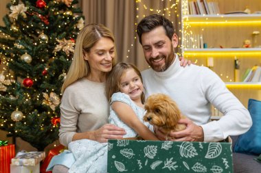 Mutlu ebeveynler, Noel Baba 'dan Noel ya da yeni yıl için bir çocuğa sürpriz bir köpek yavrusu hediye ederler. Mutluluk. Baba ve anne çocuklarına sürpriz yapıyor. Küçük kız evde bir hediye alıyor..