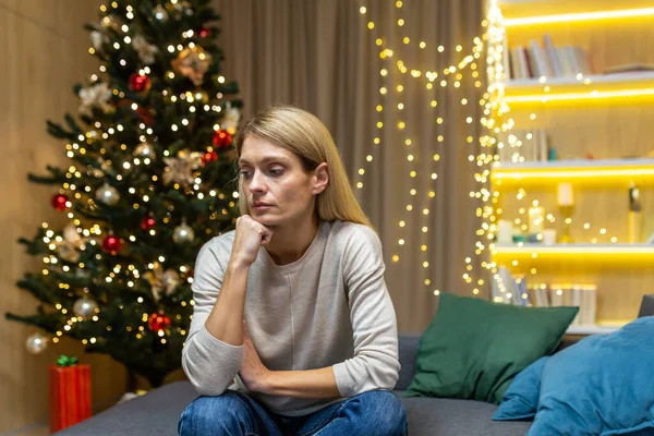 Sad woman alone at home for christmas, sitting on sofa in living room depressed on new year holidays.