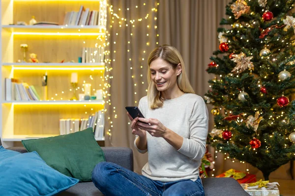 A happy young woman is holding a phone and placing an order for food delivery for a festive dinner. Sitting on sofa at home with New Year and Christmas decorations, lights and Christmas tree.
