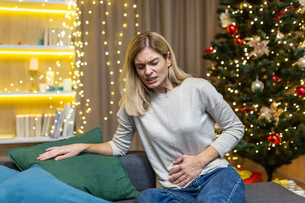 A sick woman sits at home alone, has a severe stomach ache at Christmas near the Christmas tree on New Years holidays.