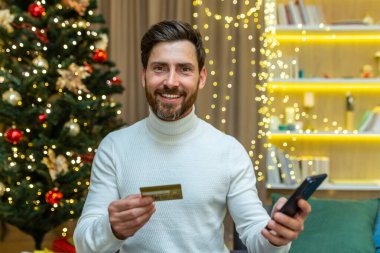 Noel adamı kameraya bakıyor ve gülümsüyor, elinde akıllı telefon ve banka kredi kartı var online alışveriş için online mağazada, yeni yıl tatilinde oturma odasında kanepede oturuyor..