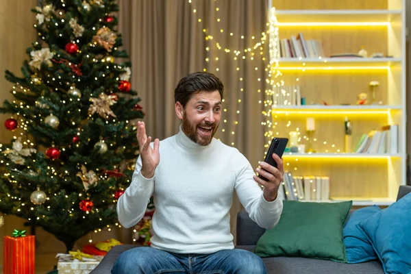 A man talks on a video call at home with a phone, sits on a sofa near a Christmas tree, records video greetings on New Years and Christmas holidays.
