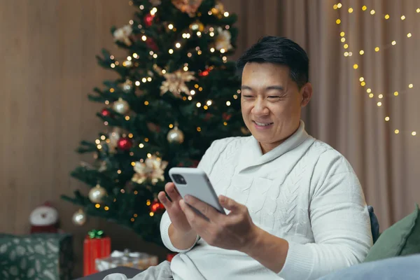 Asian at home for Christmas sitting on sofa in living room using phone, man smiling and happy holding smartphone and reading message browsing web pages online.