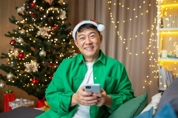 Portrait of happy man at home for Christmas, Asian man looking at camera and smiling holding smartphone, choosing gifts from friends and congratulating family online.