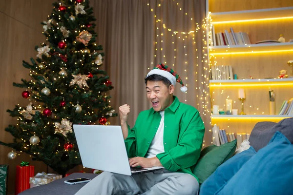 Asian man reading happy news on laptop at home for Christmas, businessman working remotely during New Year holidays sitting on sofa in living room, celebrating victory success.