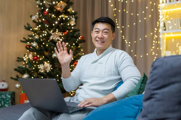Christmas man at home on sofa smiling and looking at camera, Asian man near Christmas tree with laptop holding hand up greeting gesture using laptop for remote work and new year video call.