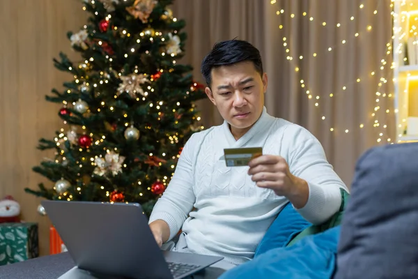Upset christmas man with laptop trying to make online purchase in online store, asian man holding bank card received error and fraud message, sitting on couch for new year.