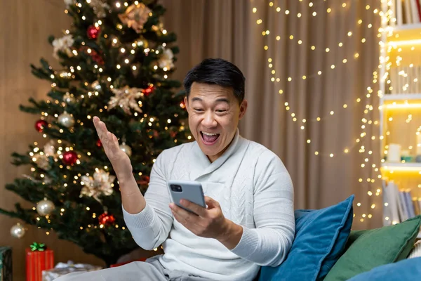 Christmas man near tree at home reading good news from phone got win and discount in online casino, Asian man holding hand up gesture of triumph sitting on sofa in living room on New Year holidays.