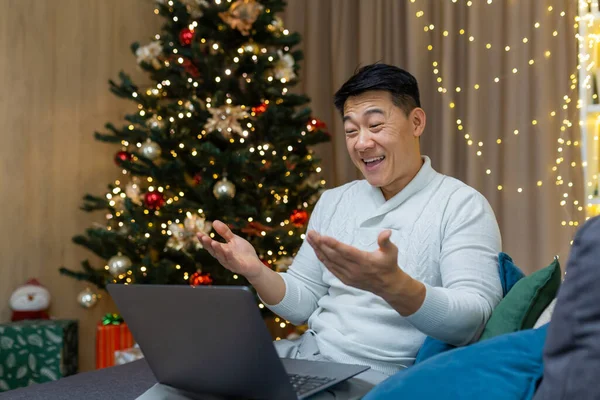 Happy christmas man making video call using laptop sitting on sofa in living room at home, asian man greeting friends online happy new year smiling and talking happily.