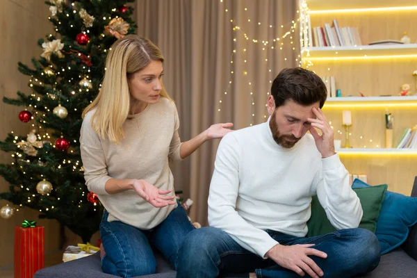 Family quarrel on Christmas New Year holiday, woman shouts and quarrels on husband, couple at home sitting on sofa near Christmas tree.