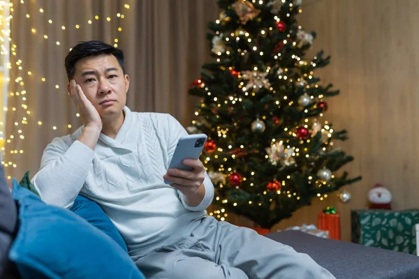 Sad and lonely young Asian man sits at home on the sofa near the Christmas tree, holds the phone in his hands. Waiting for a call, notification for the holiday.