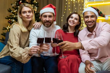 Yılbaşı için eve gelen çeşitli misafirlerin portresi, Noel 'i kutlayan arkadaşlar kameraya bakıyor ve elinde şarap kadehleri, oturma odasındaki kanepede oturuyor..