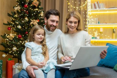Noel için mutlu bir aile evi, karı koca ve küçük kız Noel ağacının yanındaki salonda oturuyor, dizüstü bilgisayara bakıyor ve yeni yılı kutlarken gülümsüyor, internetten hediyeler seçiyor..