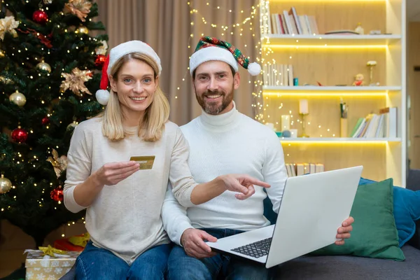 Portrait of happy family online shoppers, couple in love at home sitting on sofa near tree for Christmas holidays and new year, looking at camera and smiling, holding laptop and bank credit card.
