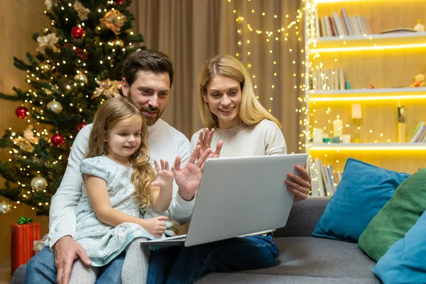Happy family greets friends via video chat, husband wife and daughter sitting on sofa at home and waving hands, online remote communication and greetings for Christmas and New Year.