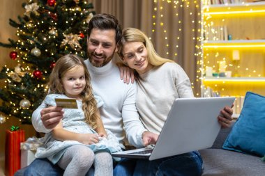 Mutlu noel aileleri online alışveriş yaparken koltukta oturuyor, karı koca, dizüstü bilgisayarlı ve banka kredi kartlı çift Noel hediyelerini Noel ağacının yanında seçerken eğleniyor..