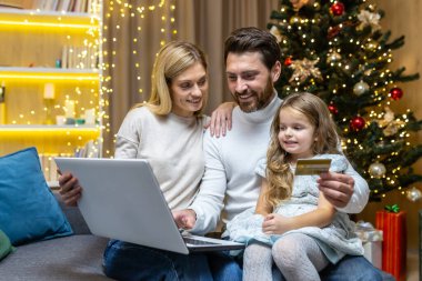 Mutlu noel aileleri online alışveriş yaparken koltukta oturuyor, karı koca, dizüstü bilgisayarlı ve banka kredi kartlı çift Noel hediyelerini Noel ağacının yanında seçerken eğleniyor..