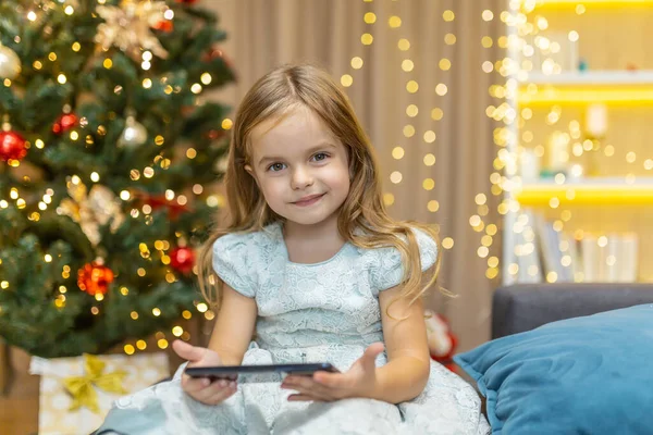 stock image Portrait of a little girl at home with a smartphone sitting on the sofa near the Christmas tree watching video content online on Christmas and New Years holidays, looking at the camera and smiling.