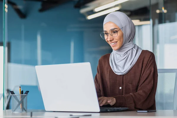 Happy and successful business woman in hijab working with laptop inside modern office building, muslim woman in glasses smiling and happy with work achievement.