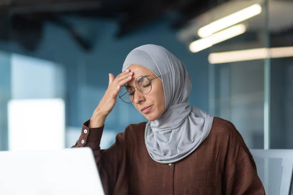 Sick woman in hijab working inside modern office building, businesswoman in glasses has severe headache, close-up photo with laptop.