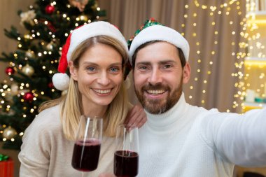 Genç ve mutlu bir çiftin şenlikli portresi. Evde Noel ağacının yanında oturuyorlar, kırmızı Noel Baba şapkaları takıyorlar. Şarap kadehleri tutuyorlar, kameraya bakıp gülümsüyorlar.