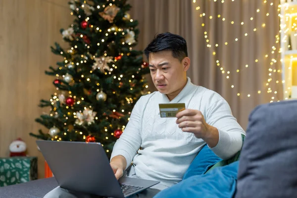 Confused and worried young handsome Asian man sitting at home and on sofa near Christmas tree. Holds laptop and credit card, cant buy gifts online, blocked card, ran out of money.