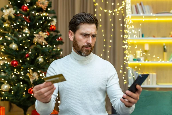 Confused and embarrassed man looks at the phone, holds a credit card in his hands. Sitting on the sofa near the Christmas tree, unable to make festive online purchases, he doesnt have enough money.