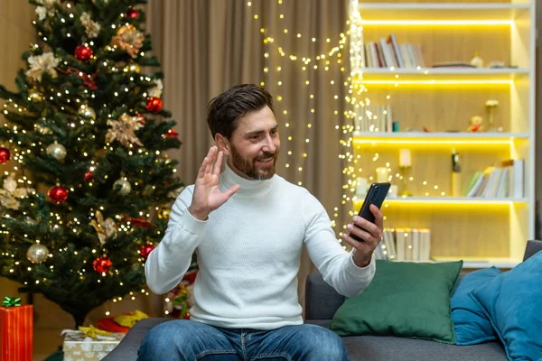 Online greetings with New Years holidays. Young handsome man at home on the sofa talking on a video call, congratulating on Christmas holidays. Holds the phone in his hands, waves his hand.