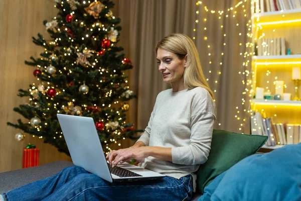 Celebration online. A young beautiful woman celebrates the New Year, Christmas online, remotely with family and friends by video call from a laptop. Sitting at home on the sofa near the Christmas tree