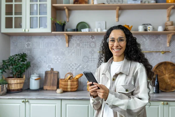 Portrait of hispanic woman at home in kitchen, woman holding smartphone smiling and looking at camera, hispanic woman wearing glasses and curly hair.
