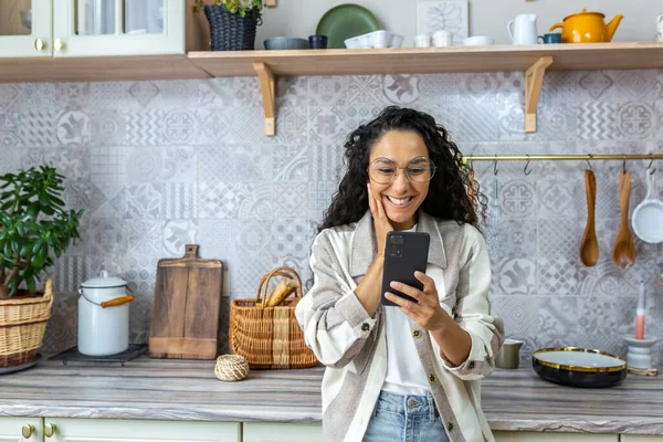 Happy Latin American woman at home reading good online news from smartphone, woman in glasses and curly hair using phone to watch online content at home in kitchen.