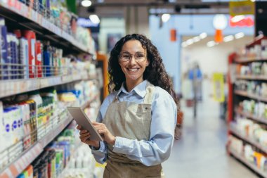 Portrait of happy and successful saleswoman, hispanic woman with curly hair smiling and looking at camera, using tablet computer to review product. clipart