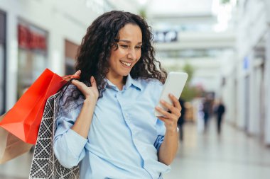Mutlu ve başarılı bir kadın süpermarkette kıyafet alışverişi yapıyor, İspanyol bir kadın elinde akıllı bir telefonla internet mesajları okuyor ve indirimli ve indirimli teklifler sunuyor.