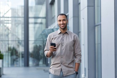 Afro-Amerikalı işadamının portresi, telefonu tutan, gülümseyen ve kameraya bakan bir adam, modern ofis binasının dışında tişörtlü bir ofis çalışanı..