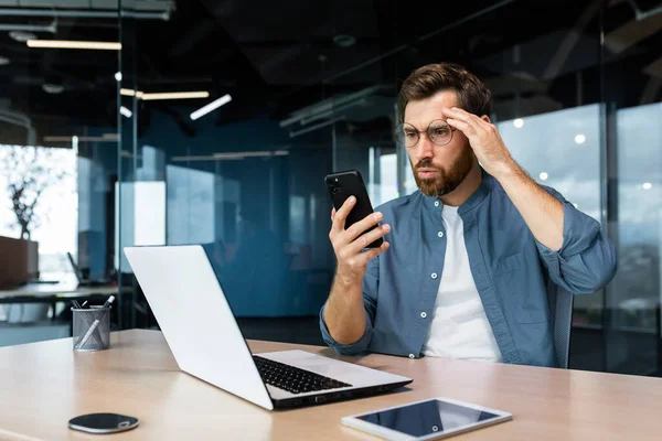 Serious mature boss working inside modern office building with laptop, businessman received bad news online, reading on smartphone confused angry and depressed man at work.