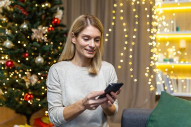 Genç bir kadın Noel tatili için ailesini ve arkadaşlarını telefonla tebrik ediyor. Elinde bir cep telefonu var, mesaj yazıyor, gülümsüyor, Noel ağacının yanındaki kanepede tek başına oturuyor..