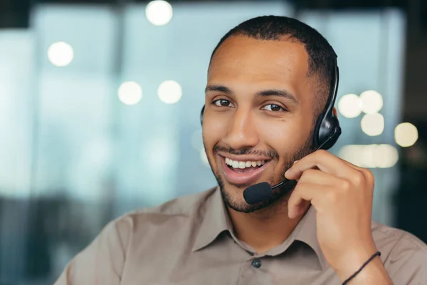 Portrait happy and smiling tech support worker with headset for video call, african american man looking at camera closeup, working inside modern office call center, online customer support service.