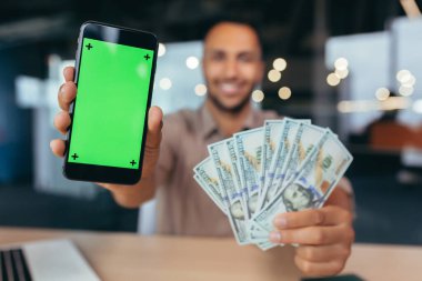 Seçici odak, modern ofis binasında çalışan iş adamı, bilgisayarlı Afro-Amerikan bir adam akıllı telefon ekralarını gösteren kameraya bakıyor ve para kazanıyor..