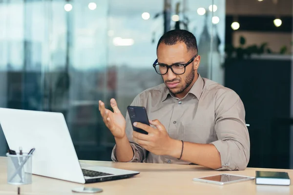 African american man received bad news notification online from phone, businessman in shirt working with laptop in middle of modern office building, man in glasses reading online letter.
