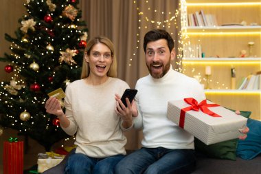 İnternetten alışveriş. Mutlu genç çift Noel ağacının yanında, ellerinde kredi kartı, telefon ve hediye tutuyorlar. Beklenmedik bir Noel paketi aldık..