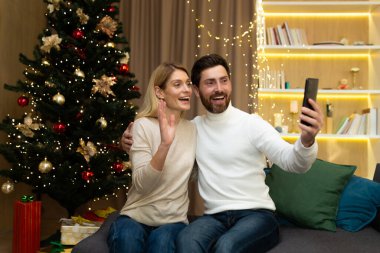 Genç bir çift, Noel ağacının yanında bir aile, ellerinde bir telefon tutuyorlar ve akrabalarıyla bir video görüşmesi yapıyorlar, Noel tatilinde arkadaşlarını tebrik ediyorlar, kameraya el sallıyorlar ve gülümsüyorlar..