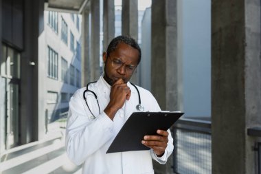 Ciddi ve olgun bir Afrikalı Amerikalı doktor modern kliniğin dışında, düşünüyor ve tedavi raporlarını ve belgeleri okuyor..
