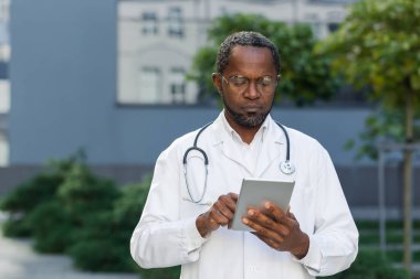 İş yerinde ciddi ve odaklanmış bir doktorun portresi, gözlüklü Afro-Amerikan bir adam tablet bilgisayarla klinik dışında çalışıyor..