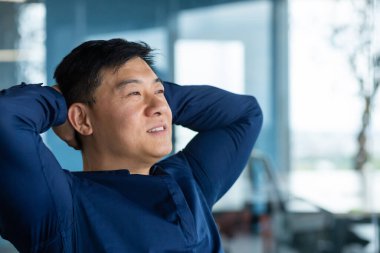 Asyalı adam pencerede dinleniyor ve hayal kuruyor, elleri başının arkasında gömlekli bir adam yan tarafa bakıyor ve gülümsüyor, gelecek planlarını düşünüyor..
