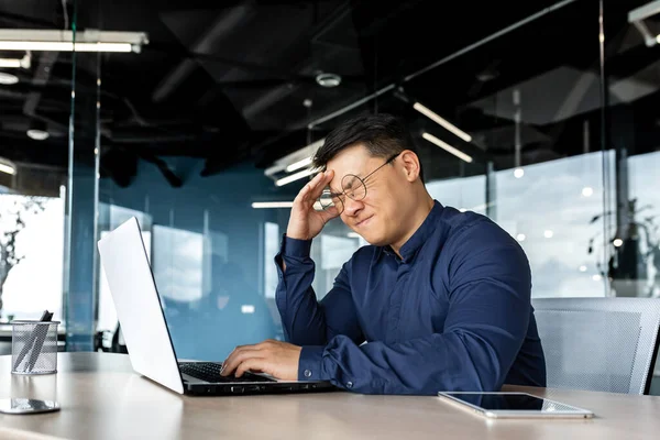 Businessman in glasses depressed, man holding his head received news of bad work result, asian man disappointed and sad working inside office with laptop.