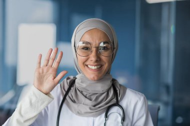 Genç, güzel bir kadının portresi, Müslüman bir kadın doktor. Bir tesettür, gözlük ve steteskopla kameraya bakıyor ve el sallıyor, gülümsüyor, selamlıyor..