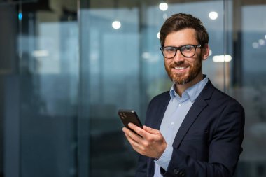 Başarılı bir iş adamı portresi, gözlüklü ve sakallı bir adam gülümseyen ve kameraya bakan, ofisteki takım elbiseli patron akıllı telefon tutuyor..
