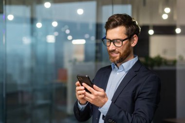 Başarılı yatırımcı ofis içinde çalışır, iş adamı takım elbiseli iş adamı pencere kenarındaki telefonu kullanır, adam gülümser ve akıllı telefondan iyi haberler okur..