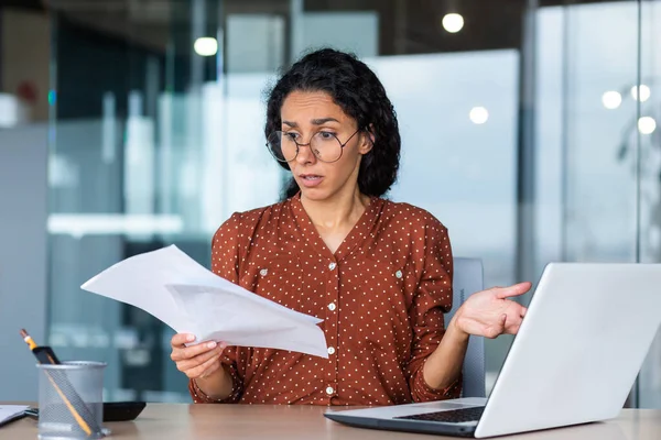 Frustrated and sad business woman holding financial report in hands, Hispanic woman sad working in office using laptop, female financier accountant unhappy with work results.